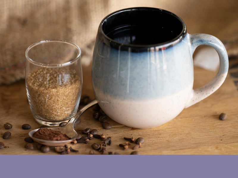 A mug of coffee next to cinnamon sticks, a spoonful of cocoa powder, and a cup of brown sugar